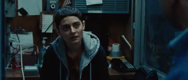 A still from the film Gazer. A short-haired young woman sits in a small, dimly lit office, looking up at an indistinct figure, with a querulous expression..