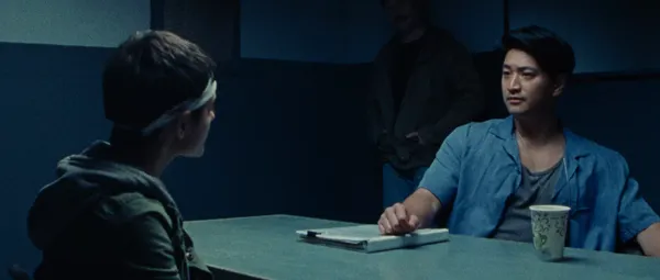 A still from the film Gazer. A young white woman with a bandage around her head sits across a table from a stern-looking East Asian man with a clipboard covered in notes in front of him. Another man stands in the corner of the small room.