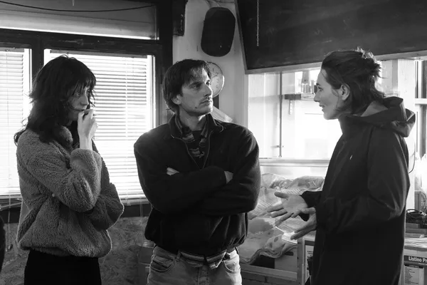 Two white women stand on either wide of a white man in a small room. The woman on the right is gesticulating to the others.