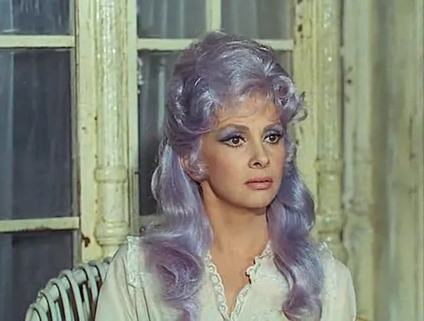 A white woman with big brown eyes. heavy lavender eye make-up and a long lavender wig.