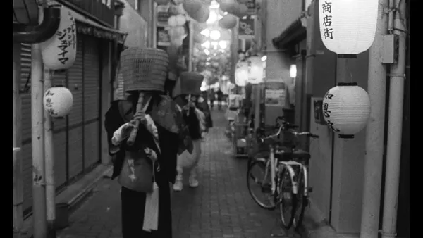 A black and white still from the film Grand Tour. A procession of robed figured with basket-like head coverings, each playing a slender woodwind instrument, moves along a narrow alleyway with bicycles at the side and balloons in the background.
