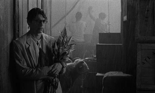 A black and white still from the film Grand Tour. A dejected-looking bespectacled white man stands on board a wooden ship as rain beats down. He is holding a bunch of lilies. Behind him is a woolly ram, and two boys are leaning over the side and waving.