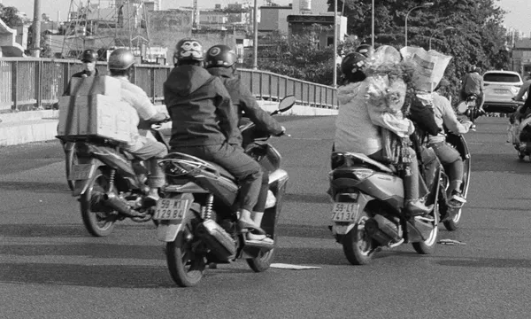 A black and white still from the film Grand Tour. A group of people on mopeds, some with passengers and one with two large boxes strapped to the back, cross a city bridge.