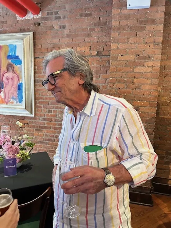 The filmmaker Griffin Dunne. A grey-haired, bespectacled older man wearing a pale striped shirt. There is an interior brick wall behind him. He looks attentive and interested.