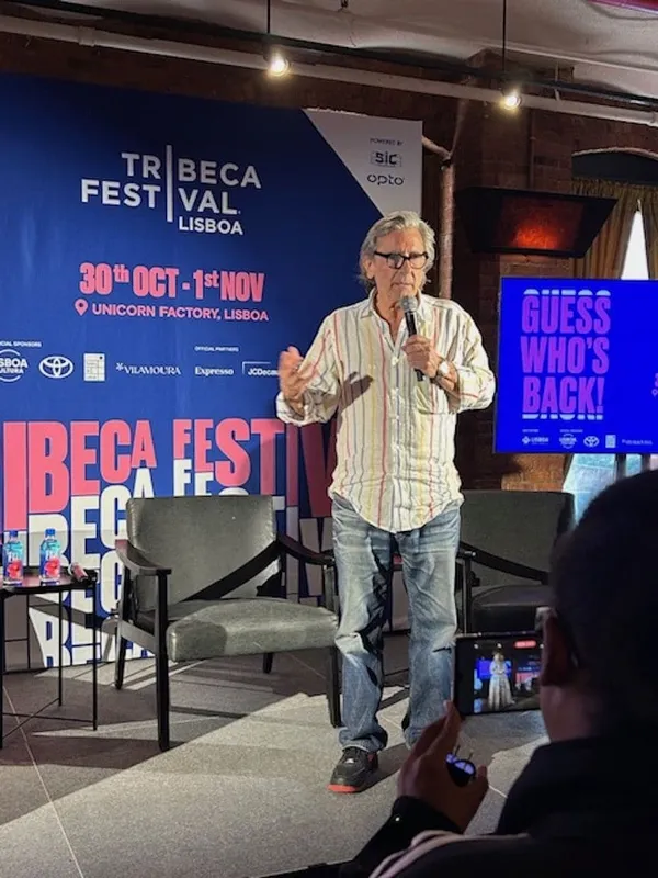 The filmmaker Griffin Dunne. A gre-haired, bespectacled older white man wearing a pale striped shirt stands on a stage surrounded by Tribeca banners, speaking into a microphone.