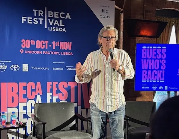 A white-haired older white man wearing faded blue jeans and a pale striped shirt stands on a stage in front of a Tribeca Festival poster, speaking into a microphone.