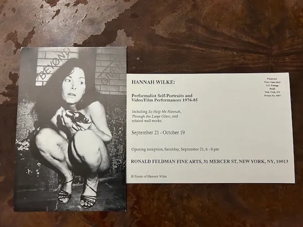A black and white photograph of a naked white woman squatting in a grimy corner, with a card beside it marked 'Hannah Wilke: performalist self portraits and video/film performances, 1976-85.
