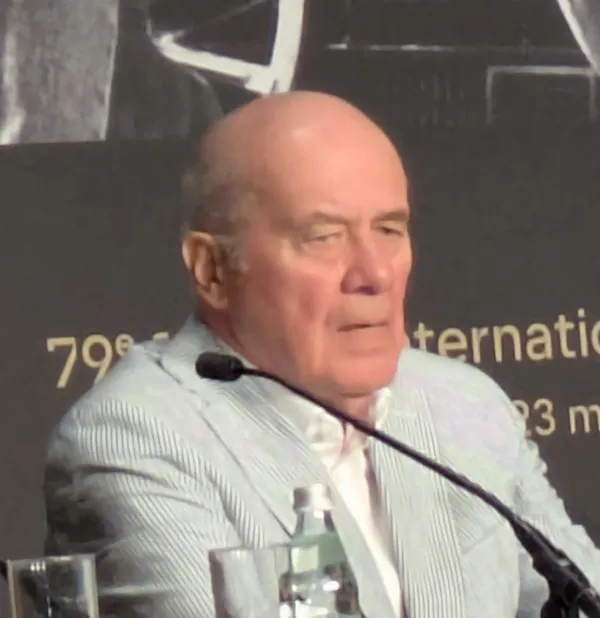 The actor Hanns Zischler. A bald white man in late miuddle age, wearing a pale grey suit with narrow white stripe detail.