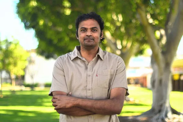 An Indian man in a pale, short-sleeved linen shirt, his arms folded, stands beneath a tree on a sunny day.
