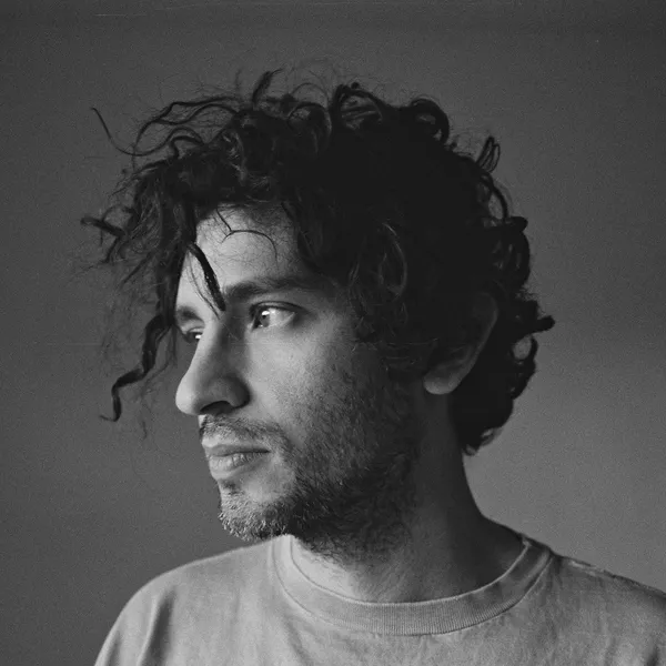 A black and white portrait of the film director Harrison Atkins. A young men with light coloured skin, a thick growth of stubble and wild, wayward black hair.
