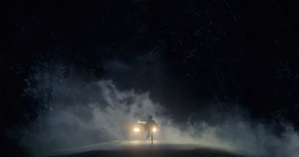 A still from the film Hellcat. A feminine figure runs along a misty road at night, beneath huge trees, followed by a vehicle.