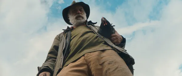 A still from the film Hen. Show from a low angle, an old white man looms large against a blue sky, holding a hen in the crook of his left arm.