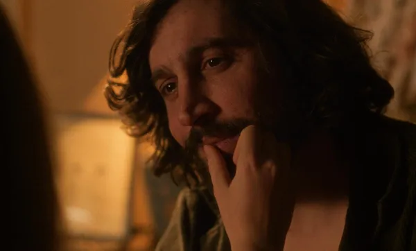 A still from the film Hotel Acropole. A slender white hand touches the face of a man with light brown skin, shoulder-length curly hair and an untidy moustache.
