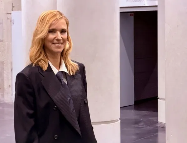 A smiling white woman with should-length blond hair, wearing a suit, stands alone near a doorway in a pillared hall.