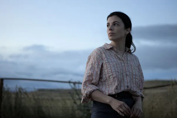 A still from the film In Vitro. A white woman with long dark hair, blue jeans and a pale checked shirt stands in a field at twilight.