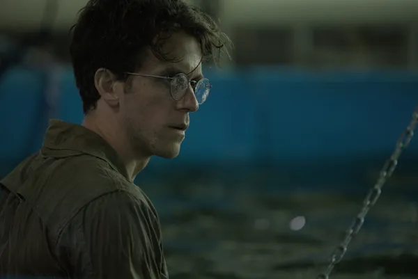 A still from the film In Vitro. A stressed-looking bespectacled white man stands in a large tub of water, with machinery in the background.