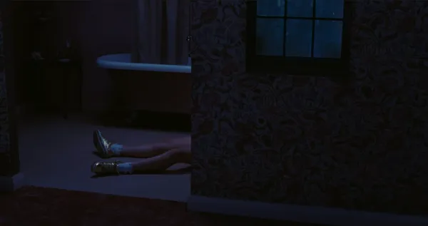A still from the film I Live Here Now. In a dimly lit house, a partial view into the bathroom reveals a white woman's legs on the floor, with white socks and gold sneakers.