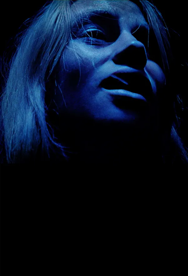 A still from the film I Live Here Now. A blond white woman's face is distorted in psychic distress, lit by blue light.