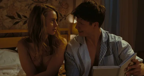 A still from the film I Live Here Now. A young blond white woman wearing just a bra and a young dark haired white man in an open striped shirt, holding a book, sit together on a bed.