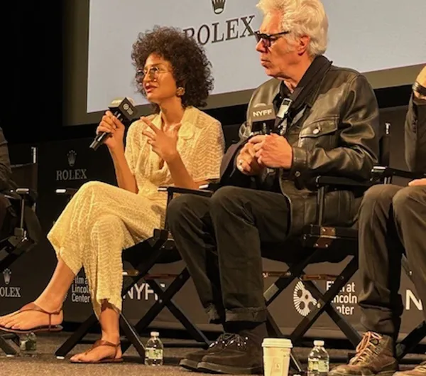 Indya Moore with Jim Jarmusch. A woman with light brown skin, big dark hair and a short-sleeved cream lace suit speaks into a microphone, beside a bleach-haired white man in a black leather jacket.