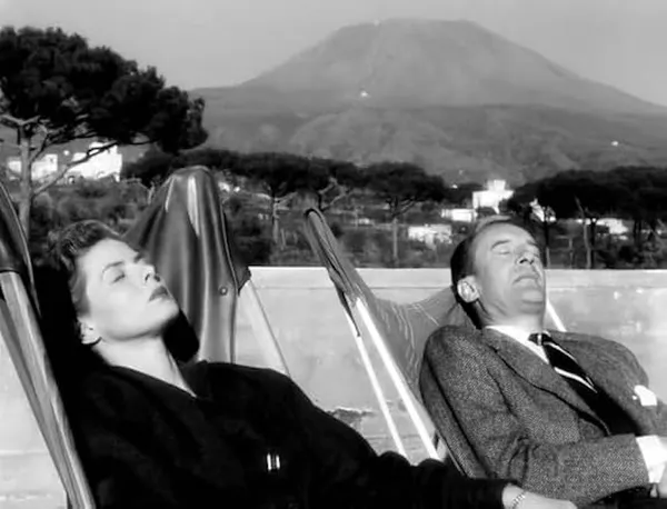 A still from the film Voyage In Italy. A fur-clad white woman and an older white man in a suit relax in deckchairs on a balcony, with the rooftops of a city below them and a volcano in the background.