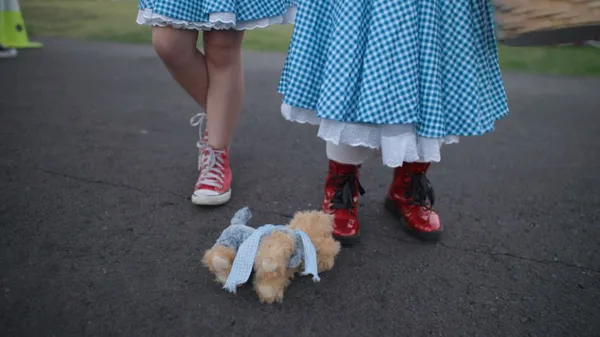 A still from the film It's Dorothy! Two children wearing blue gingham skirts are visible from the thighs down. One is wearing red PVC Doc Martens, and a small toy dog is lying beside them.