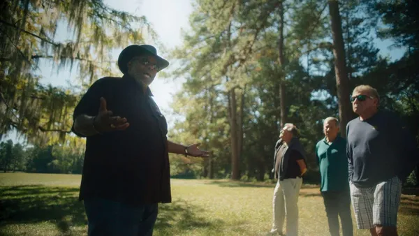 A still from the film Natchez. A Black man stands in a field hemmed in by magnolia trees, gesticulating as he talks to three older white men.