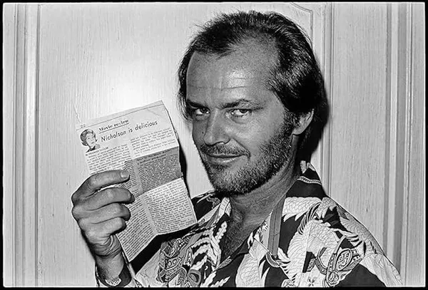 In black and white, a smiling white man - the actor Jack Nicolson - holds up a newspaper clipping entitled 'Nicholson is delicious.'