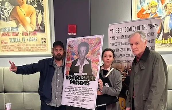 Two white men and a white woman stand in a cinema lobby, around a poster bearing the words 'Ron Delsener Presents'.