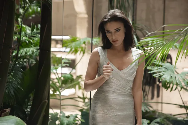 A still from the film La Gioia. An elegant white woman with dark hair and a figure-hugging silvery-white dress stands in a mirrored room full of cultivated plants.