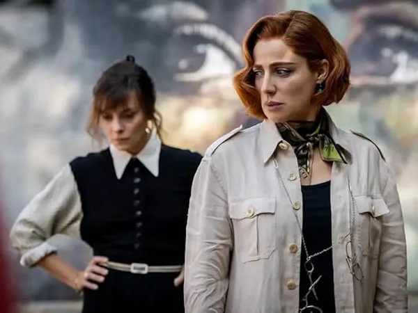 A still from the film Diamonds. Two white woman stand in front of an image of a face with huge brown eyes. One of them is wearing a black waistcoat over a white shirt; the other is wearing a cream trenchcoat and has scissors hanging from a neck chain.