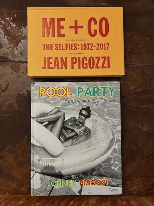 Two books on a wooden surface. A small yellow one is titled 'Me + Co: The Selfies 1972 to 2017'. The other is titled 'Pool Party' and has a black and white image of a white woman lounging on a pool inflatable. Both bear the name Jean Pigozzi.