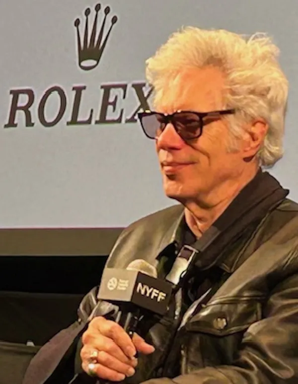 The filmmaker Jim Jarmusch. A white man with bleached hair and sunglasses talks into a microphone maked NYFF.