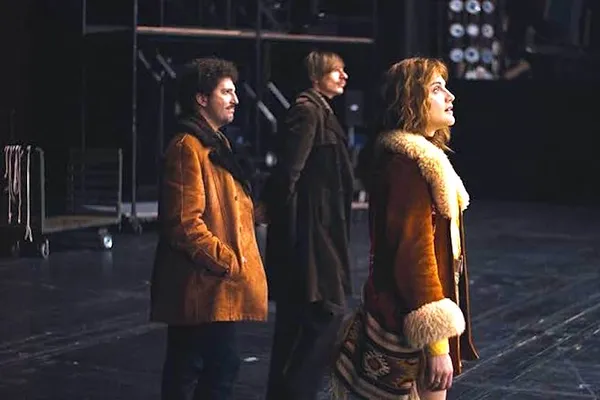 Three white people with 1970s haircuts and clothing stand in a large industrial space, all facing in the same direction.