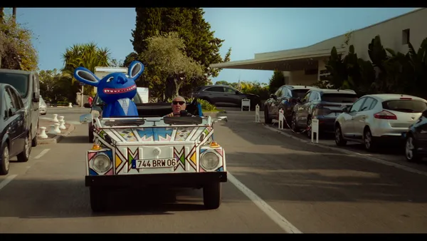 An older white man drives a boxy-looking open-top car decorated with brightly coloured patterns. In the passenger seat is a large blue grinning creature with long ears.