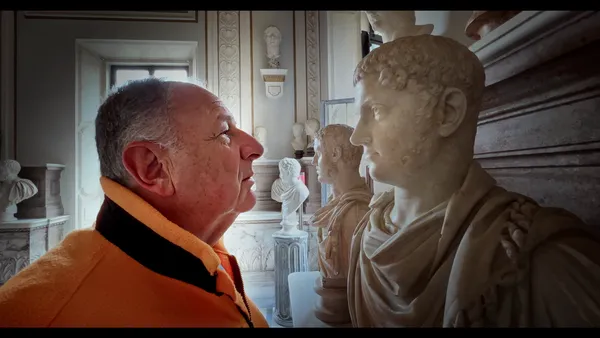 An older white man stares into the blank eyes of one of a series of marble busts of male figures.