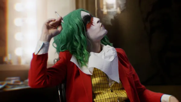 A still from the film The People's Joker. A young white woman in clown make-up, a green wig, a red jacket and a yellow checked waistcoat sites in a dressing room nonchalantly smoking a cigarette.