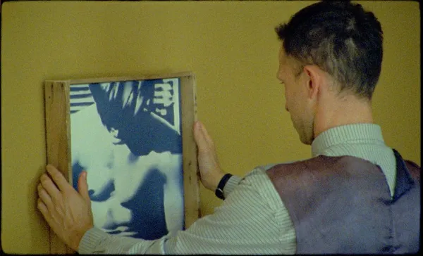 A white man positions a painting on a yellow wall.