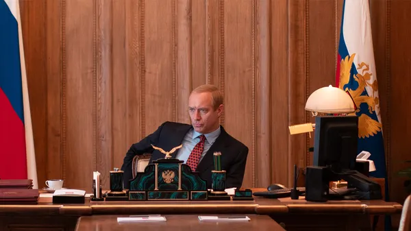 A still from the film The Wizard Of The Kremlin. A thin, white, balding politician sits behind a desk with Russian flags behind it.