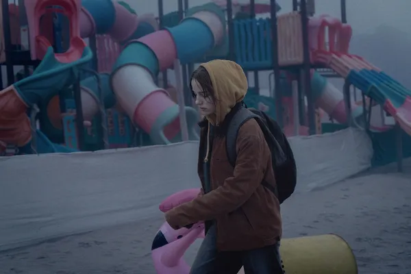 A still from the film Paternal Leave. A miserable-looking white girl walks past a colourful adventure playground on a beach, carrying an inflatable flamingo.
