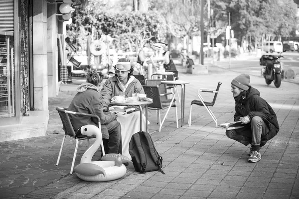 A black and white photograph of a film director instructing two cast members as they sit at an outdoor café table beside an inflatable flamingo.
