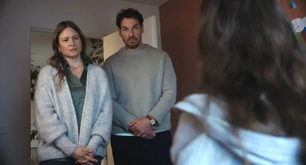 A still from the film What Marielle Knows. A girl's head and shoulders are visible from behind as a white woman and man stand in a doorway, staring at her.