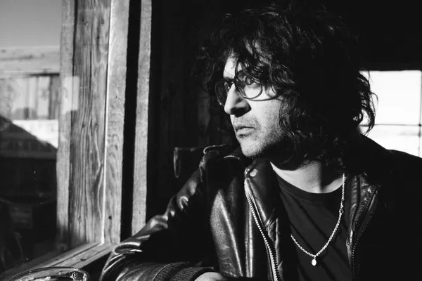 A black and white shot of the filmmaker Karan Kandhari looking out of a window. He's an Indian man with black-framed glasses, shoulder-length curly hair and a black leather jacket.