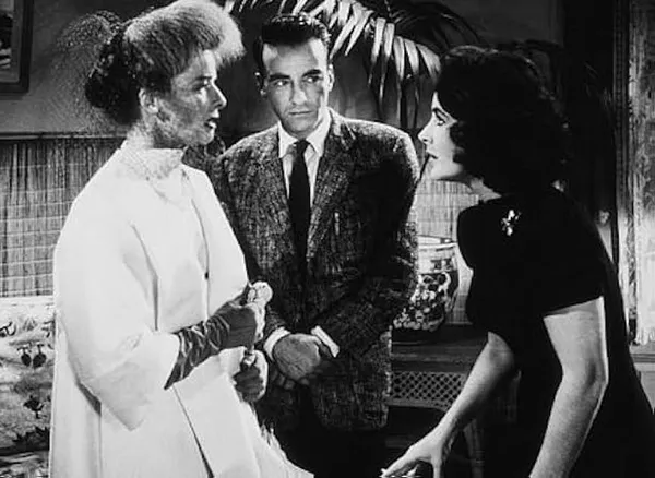 A still from the film Suddenly, Last Summer. Two glamorously dressed white women with intense expressions converse while a white man stands in the background looking slightly awkward.