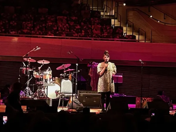 A white woman stands on a stage, holding a microphone, in front of a drum kit in a crowded auditorium.