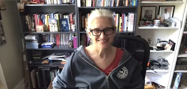 Kelly Anderson. A white-haired, bespectacled, middle-aged white woman, smiling broadly, sits in front of cluttered bookshelves.