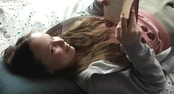 A still from the film What Marielle Knows. A smiling white girl aged about 12, with long light brown hair, lies on a bed reading a book.