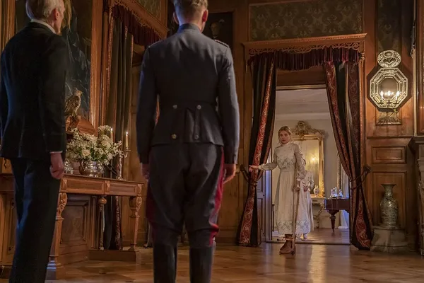 A still from the film The Kiss. An older white man in a suit and a younger white man in a military uniform stand in the hallway of an elegant Georgian home, looking at a young blond white woman who is walking with the aid of a crutch.
