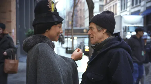A still from the film Late Fame. A Japanese woman and a white man, both dressed in winter clothes, talk in an urban street.