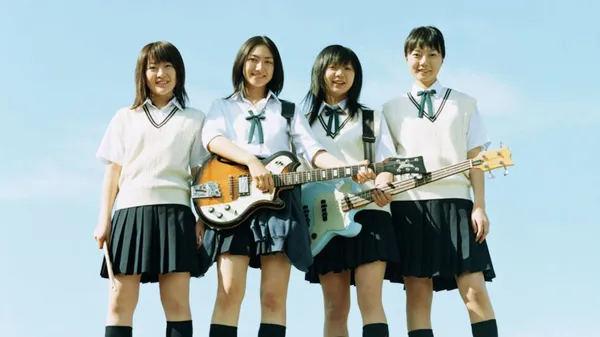 A still from the film Linda Linda Linda. Four uniformed Japanese schoolgirls stand in a row, with blue sky behind them. The one on the left id holding drumsticks and the two in the middle are holding guitars.